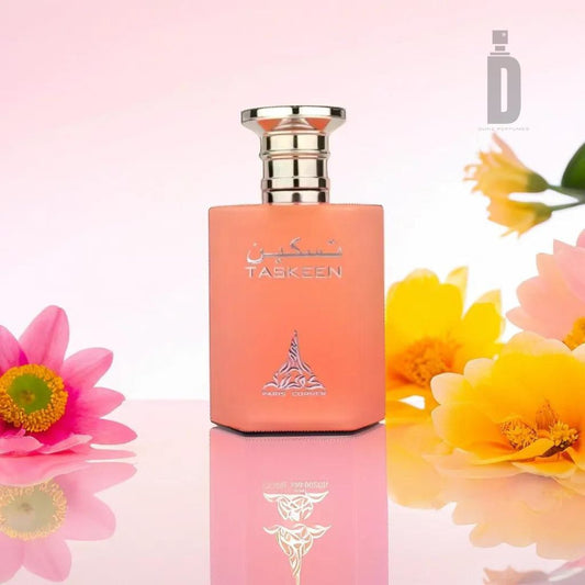 A pink and orange Taskeen 100ml EDP - Paris Corner perfume bottle with a silver cap and Arabic text stands amidst vibrant pink, yellow, and orange flowers on a soft pink gradient background. The Paris Corner logo featuring "D" is in the top right corner. The bottle's reflection is visible on the glossy surface, showcasing its luxurious fragrance.