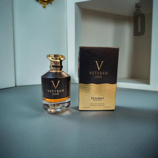 A bottle of Veteran Oud Pendora 100ml EDP - Paris Corner stands next to its matching box. The bottle features a black cap and a sleek design, while the box sports a black and gold color scheme. Both items, crafted by Paris Corner, are placed on a gray surface against a white and gray backdrop.