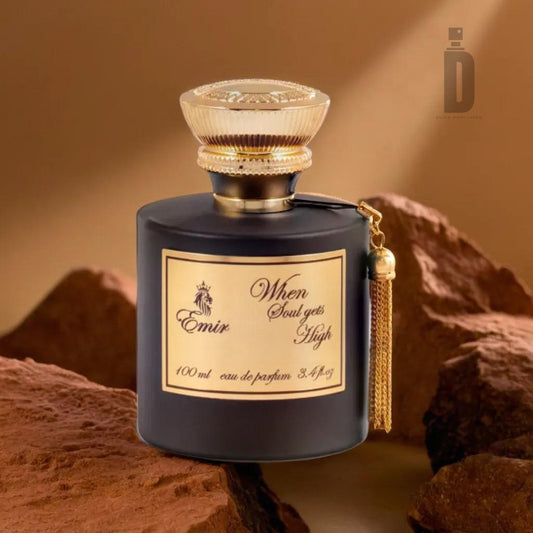A black and gold bottle of "When Soul Gets High" 100ml EDP by Paris Corner's Emir collection, adorned with a tassel, sits elegantly on a rocky surface. The bottle's cap features an intricate design, hinting at the captivating blend within. Blurred brown rocks form the background. The label prominently displays "100 ml" and "eau de parfum.