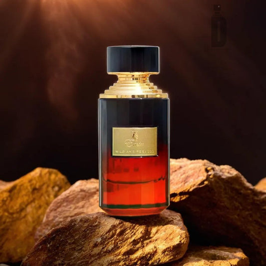 The Wild and Tobacco 75ml EDP by Paris Corner - Emir sports a luxurious fragrance bottle with a gradient design from dark at the top to amber at the bottom, featuring a gold cap and label. The bottle is elegantly placed on a backdrop of rustic rocks with a warm, dark background, evoking the rich aroma of wild tobacco.